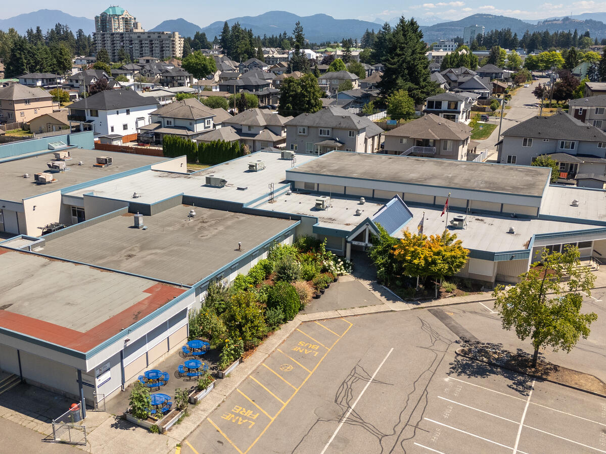Exterior image of an elementary school in the city of Abbotsford. Greenery landscape at the front entrance to the building with the city neighbourhood in the background along with some mountain range in the distance.